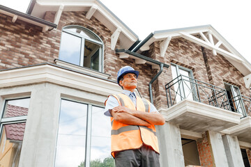 senior handsome indian worker in vest with blue helmet on head in construction process with arms crossed. on site, summer day