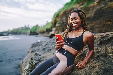 Happy dark skinned female jogger in earphones reading positive content text and smiling during sportive break at seacoast using internet connection for social networking, cheerful fit girl with mobile