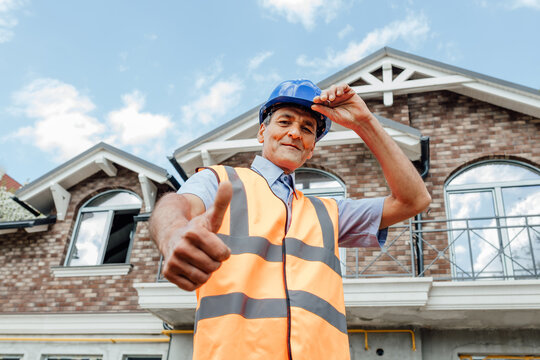 Cheerful senior male indian builder making like gesture using thumbs-up wearing work clothing vest and blue hardhat on construction site background