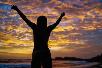Back silhouette view of satisfied fit girl celebrating sportive goals during evening time at coastline with picturesque colorful sky, carefree female athlete with casual figure feeling freedom