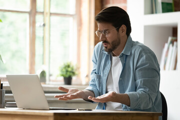 Unhappy irritated businessman in glasses looking at laptop screen, reading bad news in message, frustrated annoyed dissatisfied young man having problem with broken or discharged device, data loss