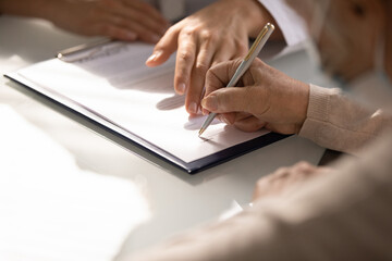 Commercial medicine. Close up of aged woman patient hand signing medical insurance contract at doctor office. Female medic show retired lady client place to put signature on healthcare coverage policy