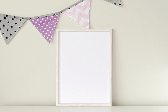 White wooden frame mockup for nursery wall art, baby girl room interior with blank frame mock up and flag bunting on wall.