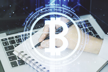 Double exposure of creative Bitcoin symbol hologram with hand writing in notepad on background with laptop. Mining and blockchain concept
