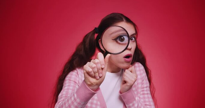 Young funny attractive caucasian girl with long braids holding a magnifying glass in front of camera and makes faces and grimaces on red backgorund. Human curiosity search concept