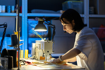 Creating new fashionable collection and garment industry concept. Young female clothes designer sew on sewing machine sitting at workplace in fashion workshop. Small business owner or tailoring hobby