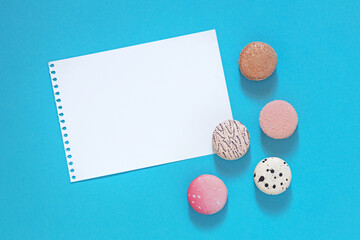 top view of sweet colored french meringue pastries macarons, white blank sheet of paper on blue background,