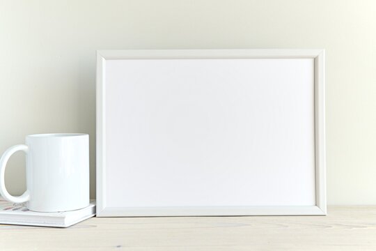 White empty horizontal frame mockup for wall art on white shelf, minimal clean interior, blank frame mock up, coffee mug, books.