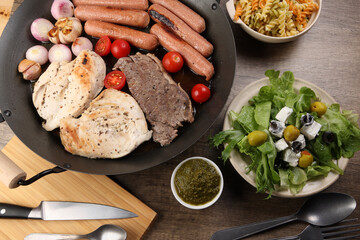 Mix meet chicken beef steak sausage tomato onion garlic fry grill oil metal fry pan colourful pasta green salad with cheese olive mint basil pesto sauce board knife fork spoon rustic wooden table