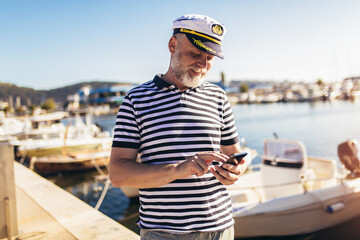 Mature man standing near the sea dressed in a sailor's shirt and hat using smart phone - obrazy, fototapety, plakaty