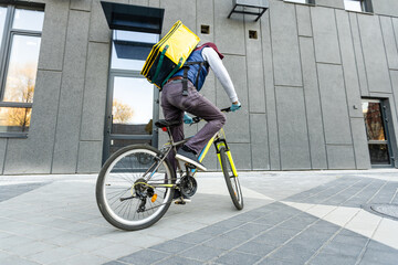 Young courier in medical mask delivering food with yellow thermal backpack, riding a bicycle in the city. Food delivery service concept