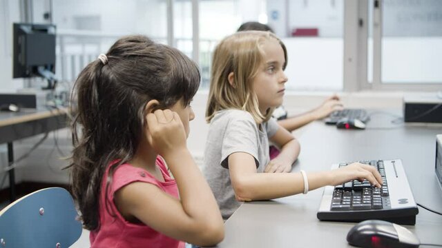 Multiethnic little girls typing on keyboard together. Schoolgirls sitting at table during computer science lesson. Blonde girl explaining task to Hispanic classmate. Friendship, informatics concept.