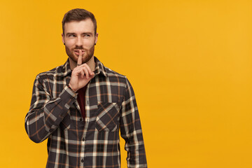 Portrait of cunning, adult male with brunette hair and bristle. Wearing checkered shirt and accessories. Shows silence sign. Watching to the right at copy space, isolated over yellow background