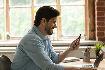 Side view smiling businessman in glasses looking at phone screen, happy young man reading good news in message, browsing apps, surfing internet, chatting in social networks, sitting at desk