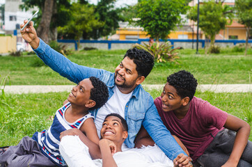 Dominican Latino origin man celebrating father's day with his children in the park, dark-skinned family having fun, children hugging their father taking a selfie sitting in the park