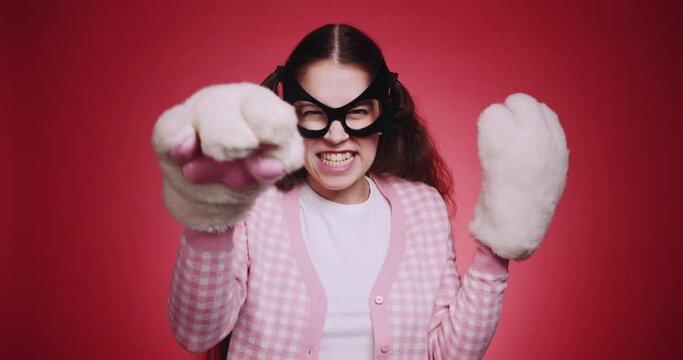 Cute girl in a cat mask and pink clothes with plush fluffy paws flirts with cat gestures in front of the camera on red background. Catwoman funny cosplay masquerade
