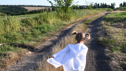 Defocus silhouette of little girl back run by country road at rural landscape in summer. Blurry white cape and blonde ponytails fly in wind. Serenity of childhood