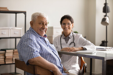Fototapeta premium My attending physician. Portrait of satisfied elderly male patient visiting doctor sit on chair in office at clinic look at camera with happy smile. Young lady therapist consult old man at workplace
