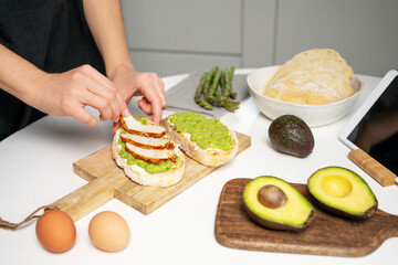 Instruction, step 5. Woman hand holding roasted chicken slice and making avocado toast with cream cheese. Fresh bread, vegetables, eggs and tablet on white table. Delicious healthy breakfast recipe.