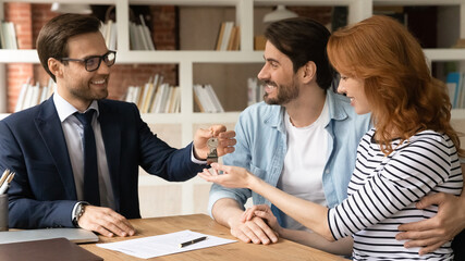 Happy millennial couple receiving keys from realtor, purchasing real estate, making successful deal at meeting, smiling man and woman buying first own dwelling, new apartment, mortgage or rent