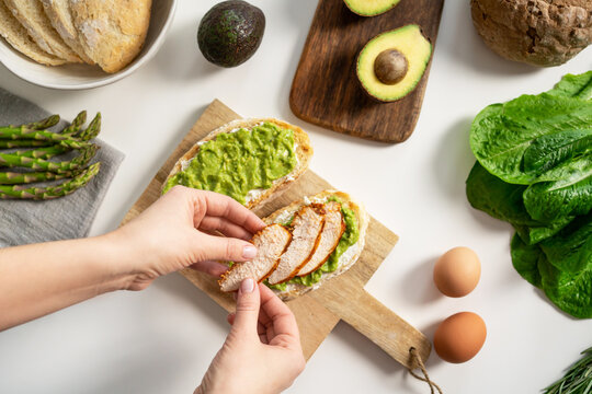 Instruction, Step 4. Female Hand Holding Roasted Chicken Slice And Cooking Sandwich With Cream Cheese And Mashed Avocado. Fresh Bread, Vegetables, Eggs And Salad On White Background. Tasty Breakfast.