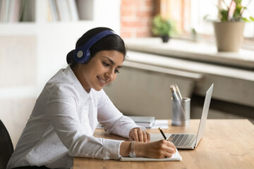 Smiling interested Indian woman in headphones writing taking notes, student using laptop, studying...