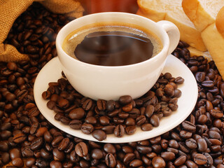 A  white cup of hot aromatic coffee near a sack of coffee beans with bread on the desk in the room