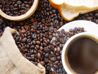 Sack of coffee beans are near a cup of hot dark coffee and a bowl of coffee beans and some breads