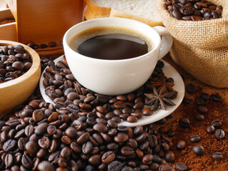 A cup of aroma hot coffee in white cup for drink in this morning with coffee powder and coffee beans in sack and coffee beans in wooden bowl and some breads