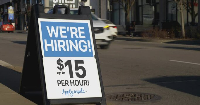 A "We're Hiring!" sandwich board sign outside of a business on a city sidewalk. Sign shows $15 per hour wage. Sign customizable upon request.	