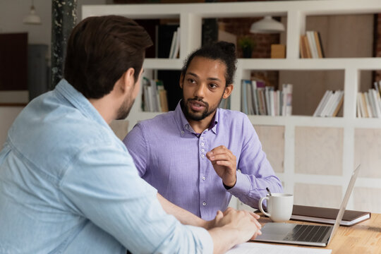 Diverse colleagues working on project together, using laptop, sharing ideas or discussing strategy, brainstorming, serious confident African American mentor coach training new employee, intern