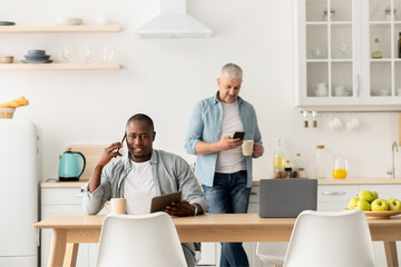 Busy morning. Mature african american guy working on tablet and talking on phone at table