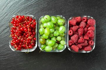 Assorted berries of raspberries, gooseberries and red currants in glass bowls on a black table.