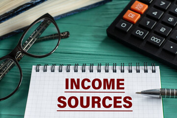 INCOME SOURCES - words in a notepad on a wooden green background with a calculator, pen and glasses