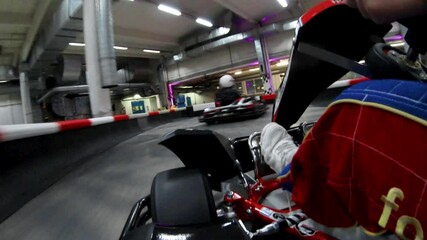 Two kart driver wearing helmets and racing suits drive over an indoor track and quickly around tight curves - on board camera on second cart