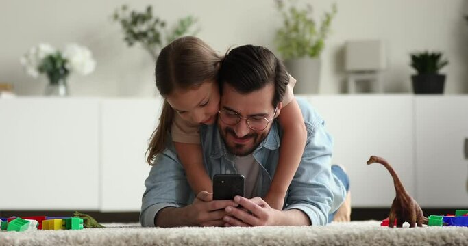 Happy young handsome father in eyeglasses ling on floor carpet with playful small child daughter on back, showing funny mobile applications, playing games, web surfing online, tech addiction concept.