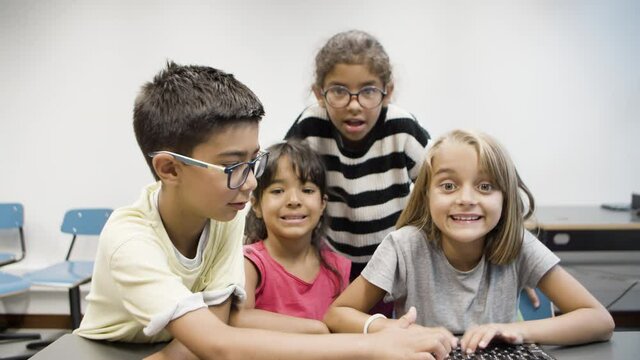 Multiethnic children having fun during computer science lesson. Smiling blonde girl and boy in eyeglasses typing on keyboard. Their classmates looking at monitor. Informatics, friendship concept.