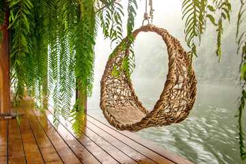 Hanging rattan chair with cushion near tropical river with green plants