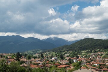 Fototapeta na wymiar Panoramic natural landscape, town with old typical traditional houses, red roofs, against green mountain hills. High dynamic contrast, dramatic clouds. Bulgaria, Kalofer, Balkan Mountains, Botev peak.