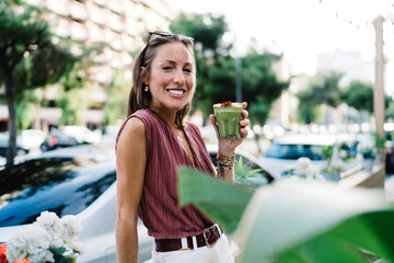 Half length portrait of cheerful caucasian woman in trendy wear standing outdoors holding glass...