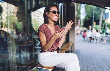Beautiful female blogger surprised with success of publication gesture emotionally holding mobile phone, smiling caucasian woman in trendy clothes and sunglasses laughing feeling confused from message