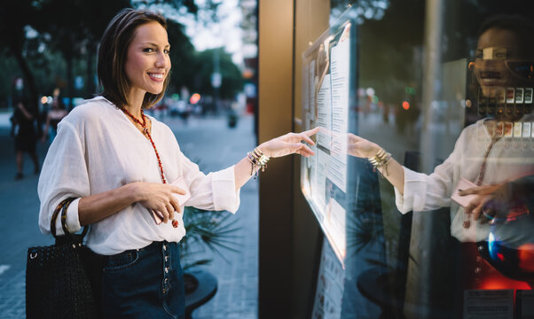 Half length of cheerful tourist smiling at camera near panel while tapping sensory for online ticketing, happy female touching screen for self consulting at electronic system using advanced display