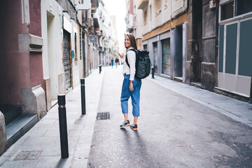 Full length portrait of cheerful woman walking at city street and smiling at camera, happy female tourist with smartphone gadget using roaming internet for searching location of quarter in town