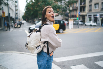 Amazed female tourist with backpack standing at urban setting and feeling shocked during international vacations for getaway, excited Caucasian woman with open mouth looking away and impressing
