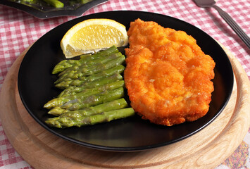 Piatto nero con cotoletta di pollo fatta in casa e asparagi bolliti e conditi con olio sale e fetta di limone.