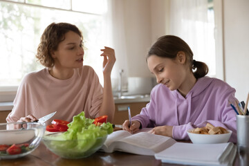 Happy mother helping young teen child daughter with learning, preparing assignment, school task or homework while cooking healthy vegetarian salad for dinner in kitchen, casual daily routine concept.
