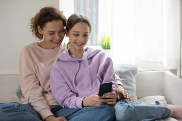 Smiling happy different female generation family resting on comfortable sofa, using software smartphone applications, playing mobile game, blogging, web surfing online, watching funny photo video.