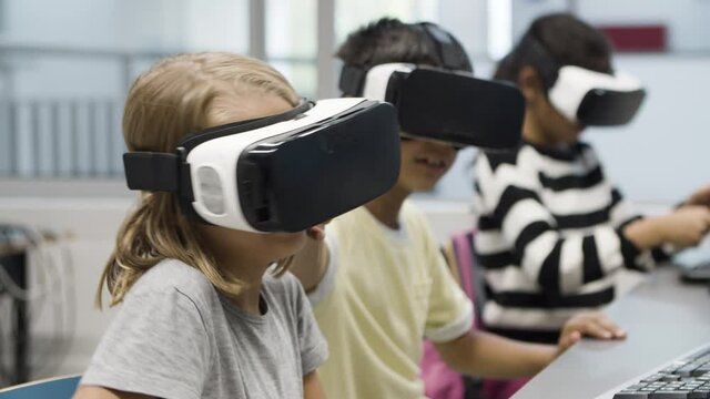 Close-up of schoolgirl wearing VR headset. Smiling blonde girl sitting at desk, imagining that she eating ice cream, looking around. VR technology in education concept.