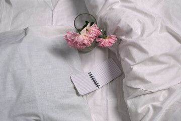 Delicate pink roses, notebook on a white unmade bed.