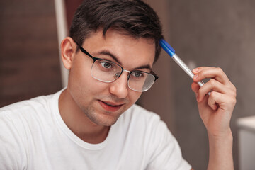 A pensive young man rubs his head with a ballpoint pen. Close-up portrait